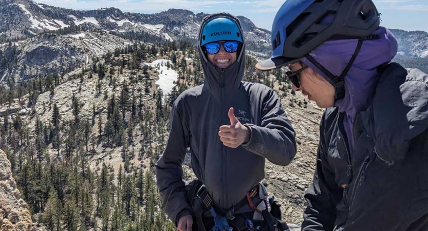 Two people wearing helmets stand at a high elevation, above snow dotted mountains. one person gives the camera a thumbs up. 